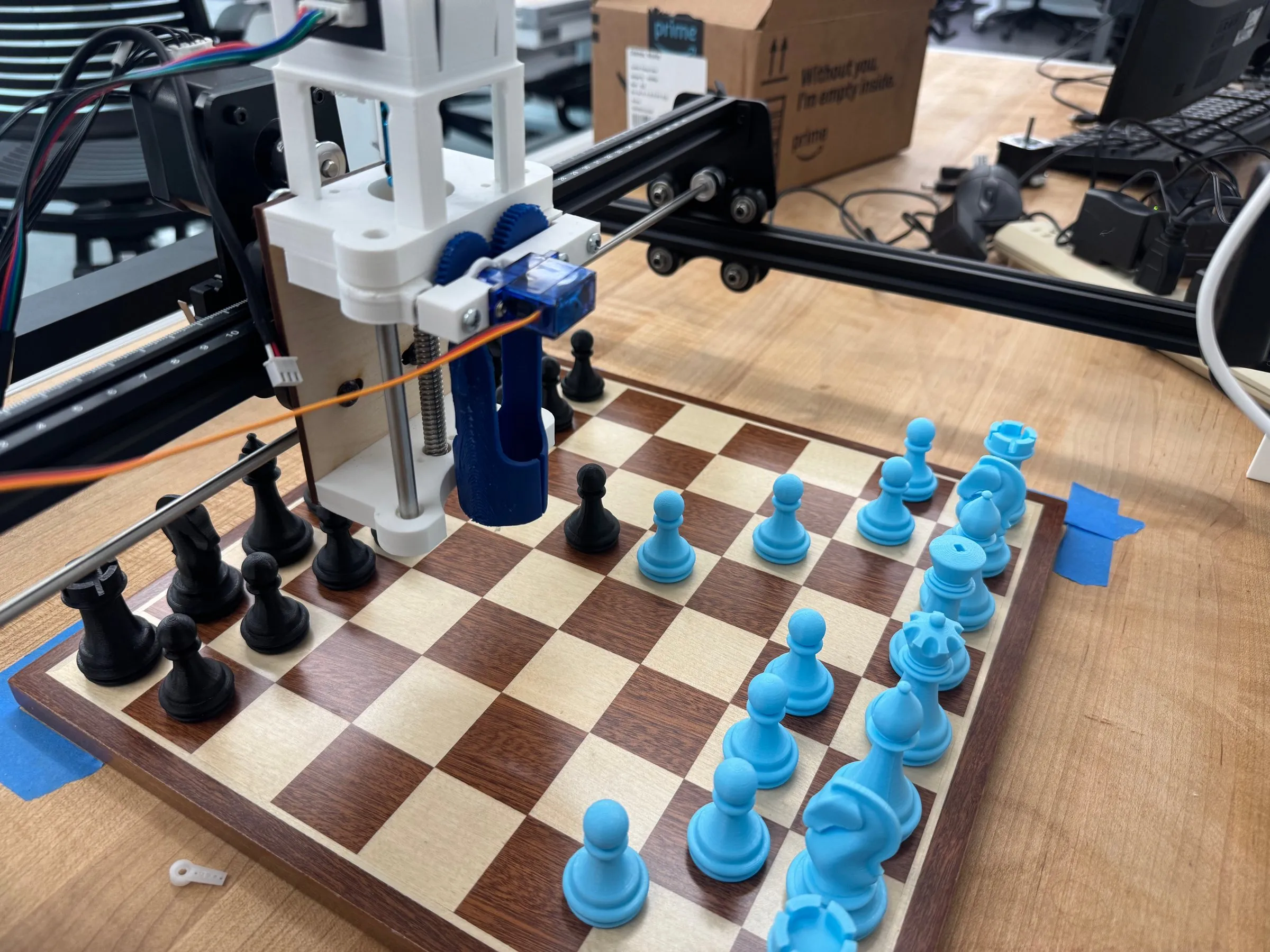 A gantry-mounted gripper hovering over a wooden chessboard set with black and blue 3D-printed pieces.