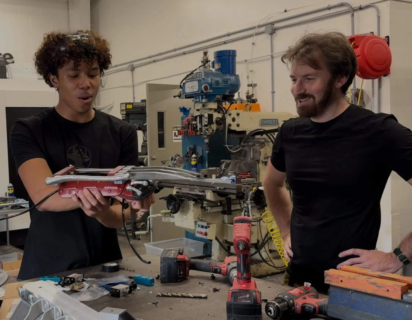 Holding the pneumatic Wolverine claws at a milling machine in the Hacksmith shop as a colleague looks on.