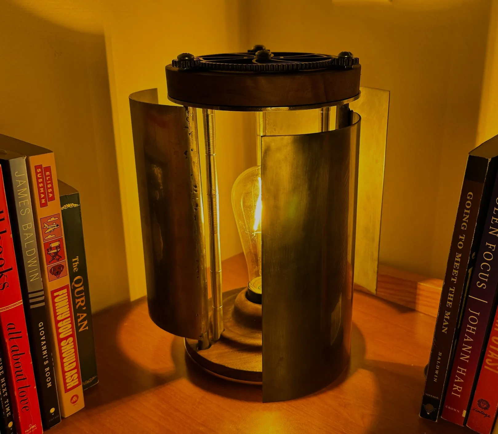 A lamp with a geared top ring and curved metal panels glowing warmly around an Edison bulb, between rows of books.