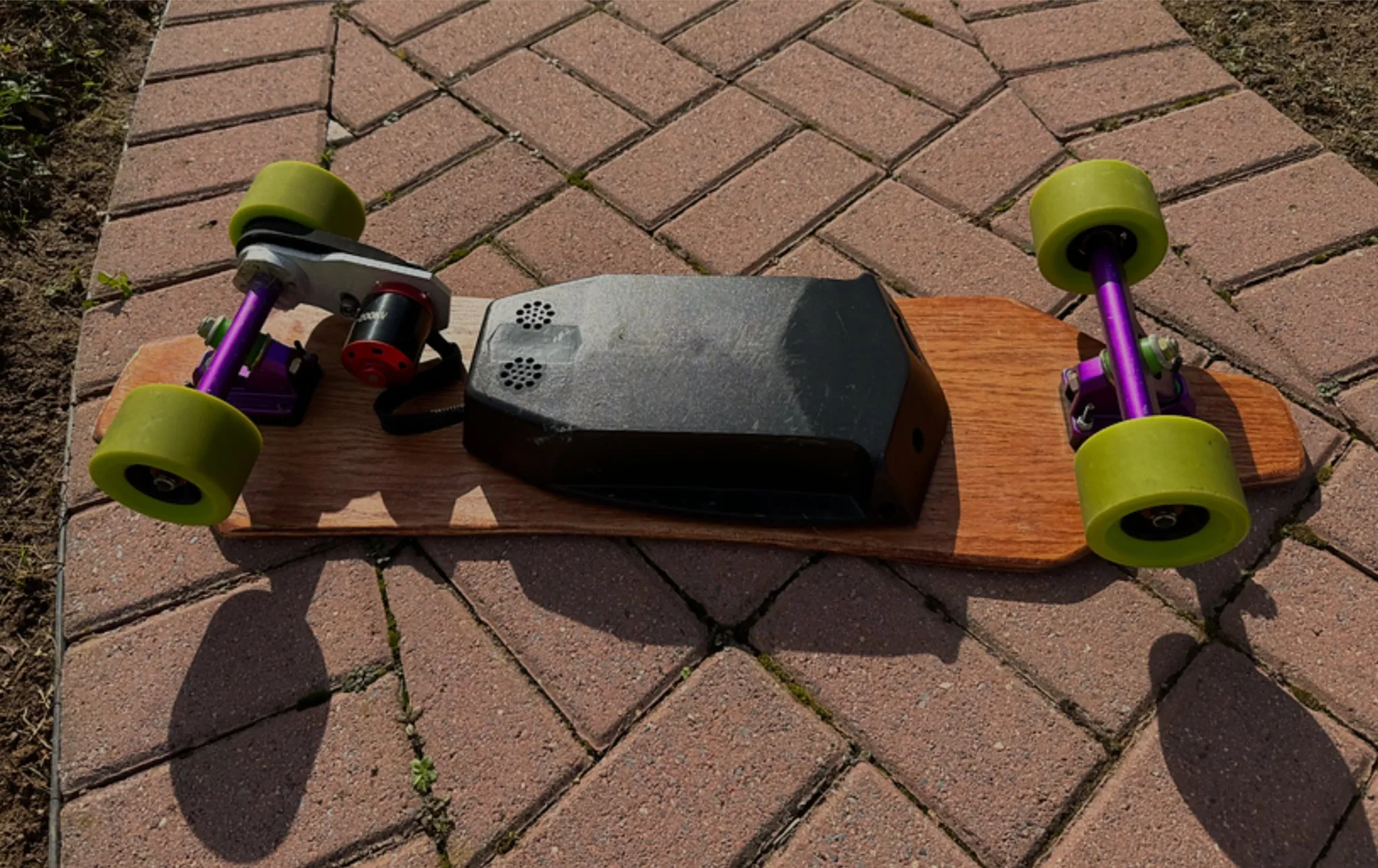 An electric skateboard with green wheels, purple trucks and a black electronics enclosure, resting on brick pavers.