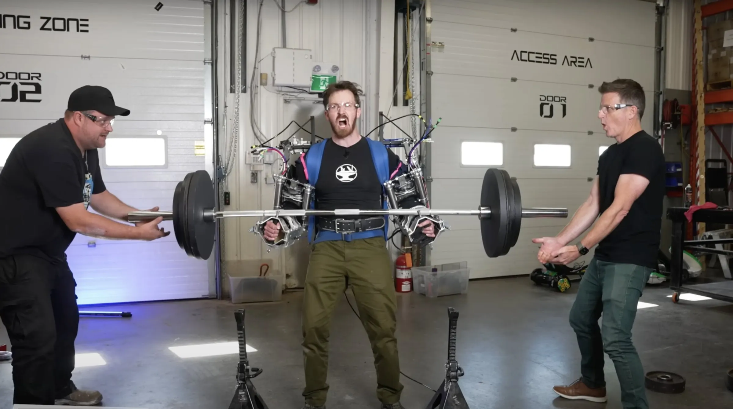 The exoskeleton deadlifting a loaded barbell in the Hacksmith shop while two engineers spot.